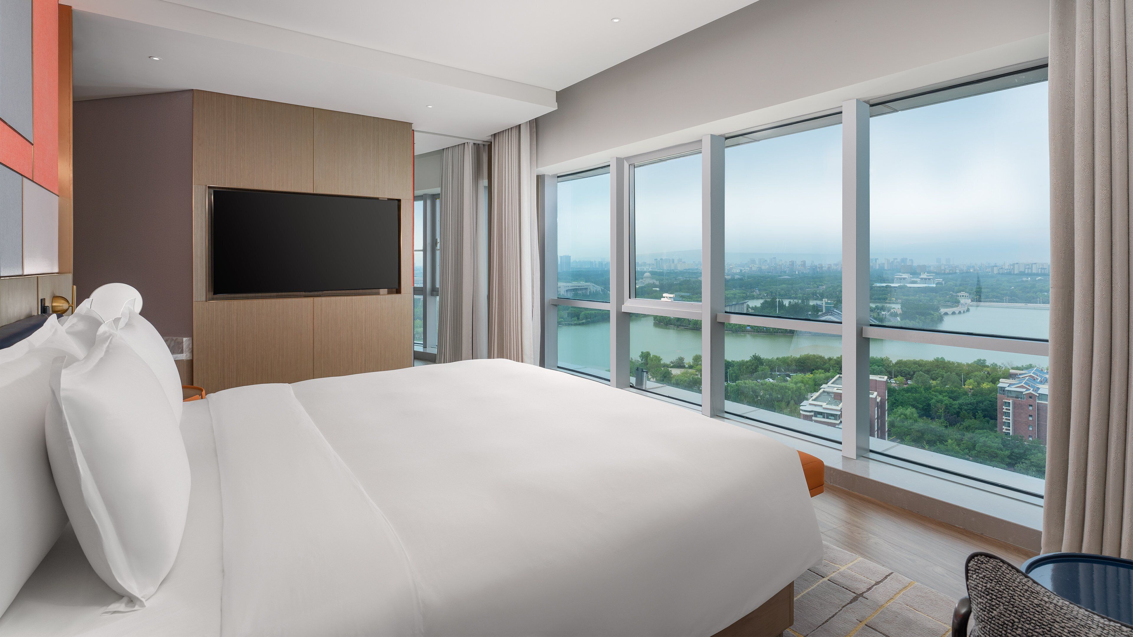A modern hotel room with a large bed, a flat-screen TV, and a city view through large windows.