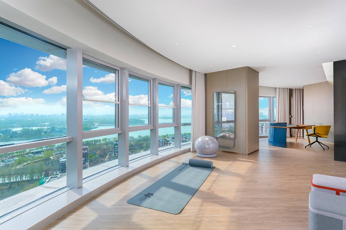 A spacious room with large windows offering a city view, a yoga mat, exercise ball, and a desk with chair.