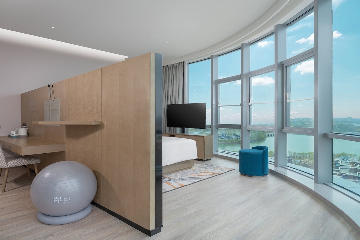 Una habitación de hotel moderna con un ventanal grande, un escritorio, una silla, un televisor y una pelota de ejercicios.