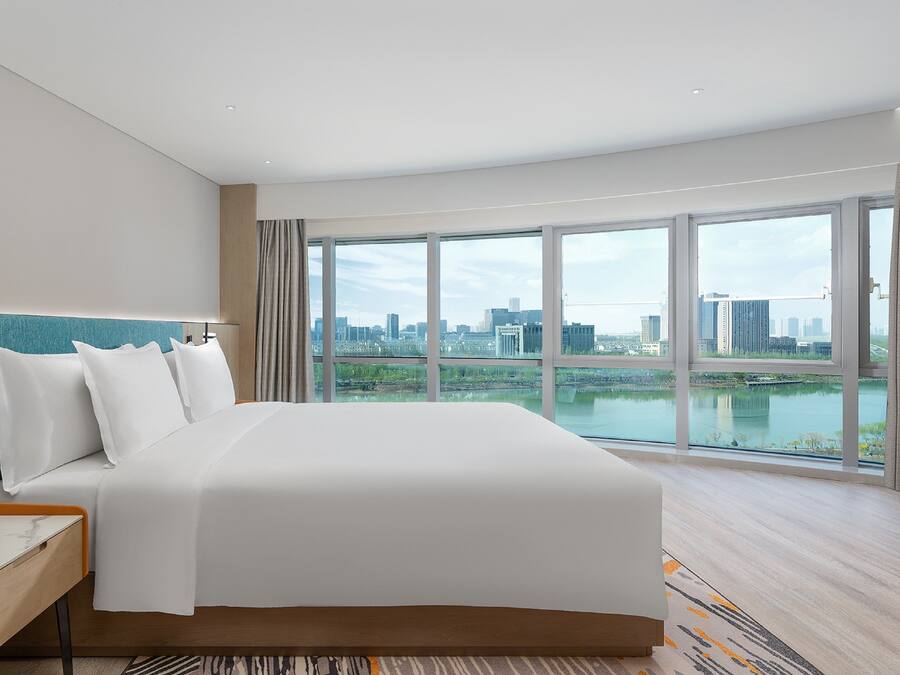 A modern hotel room with a large bed, a bedside table with a lamp, a TV, and a view of a cityscape and river.