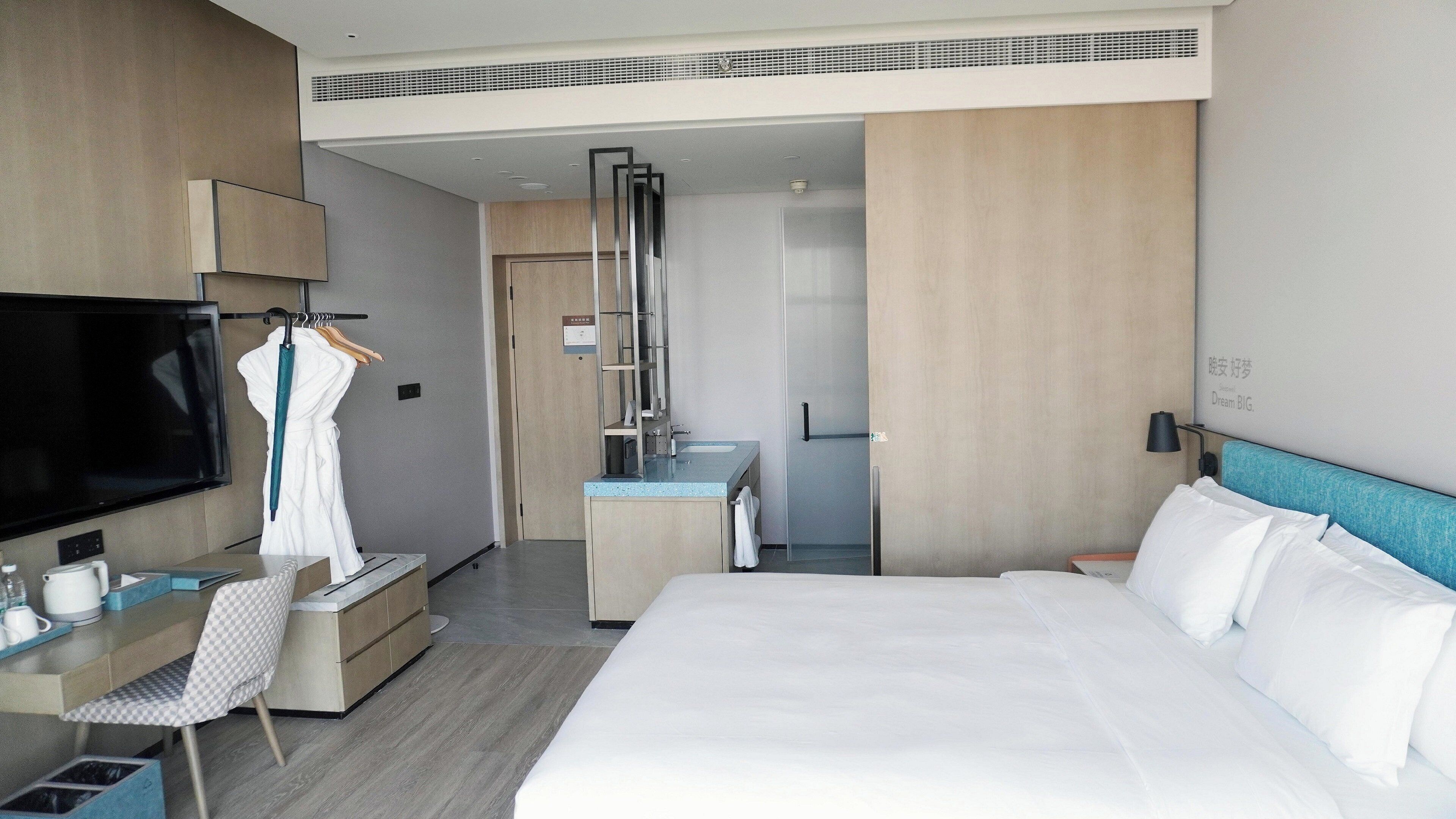 A hotel room with a bed, a desk with a chair, a TV, a wardrobe, and a bathroom with a shower.