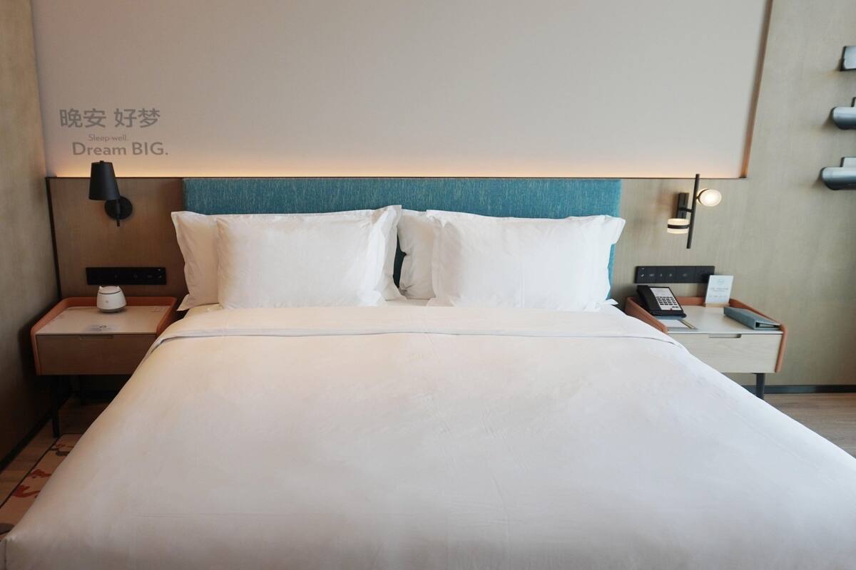 A hotel room with a large bed, a nightstand with a lamp, a bedside table with a phone, and a wall-mounted shelf.