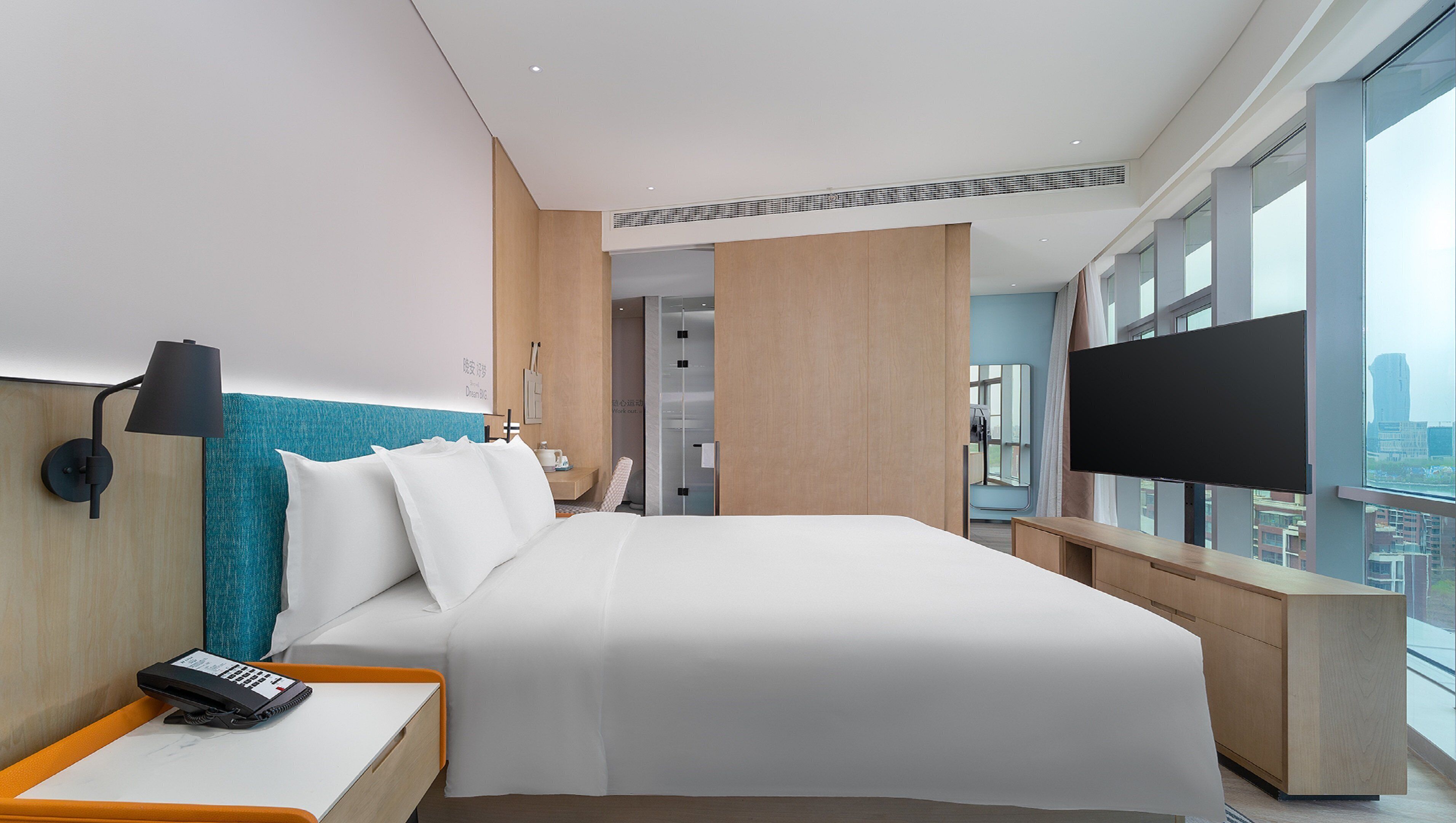 A modern hotel room with a large bed, a desk with a lamp, a TV, and a city view.