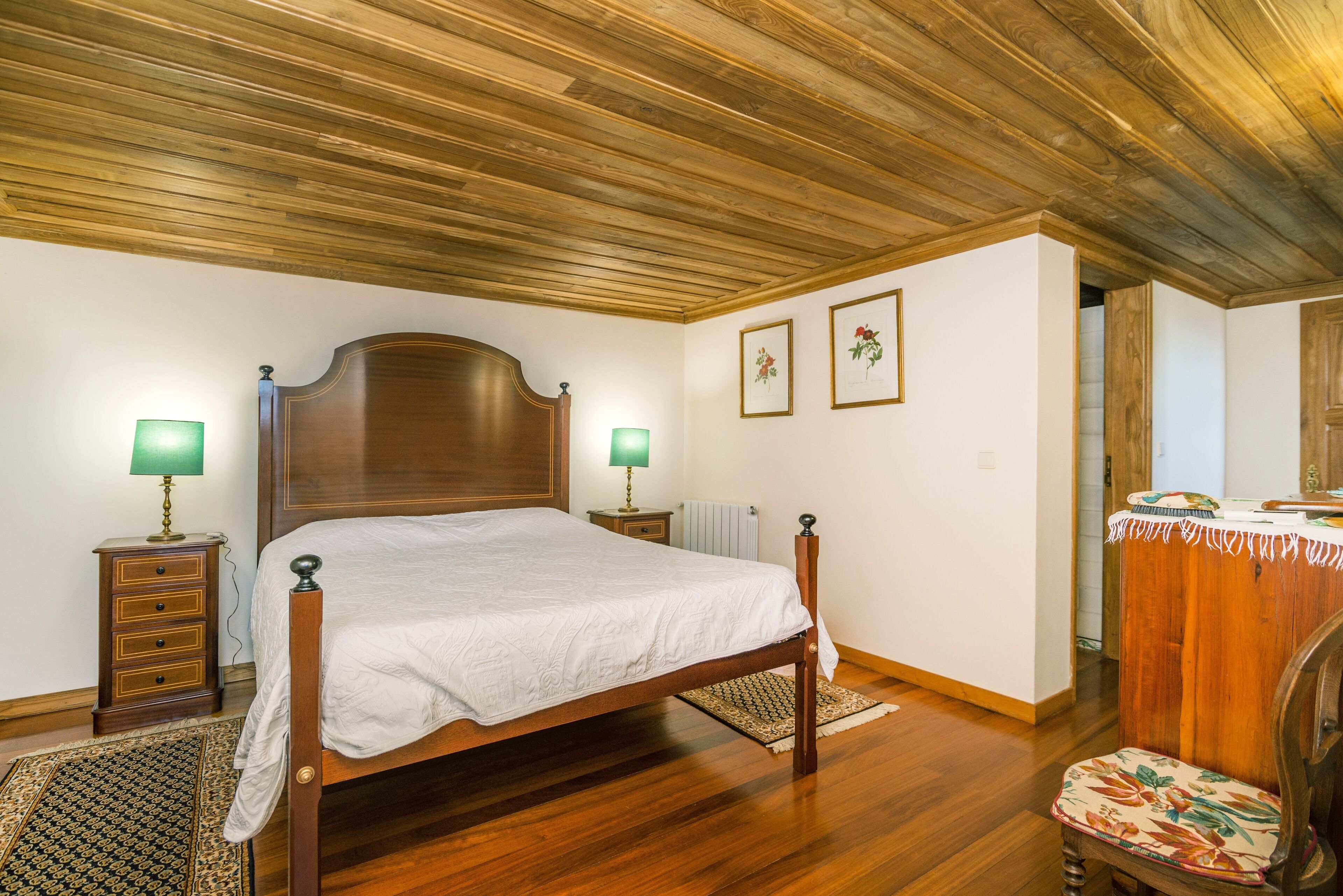 Een slaapkamer met een houten bed, twee nachtkastjes met lampen, een houten hoofdbord, een houten dressoir en een fauteuil met dessin.