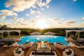 Una piscina con sillas de descanso y sombrillas, rodeada por una pérgola blanca, con vistas a la puesta de sol sobre un paisaje.