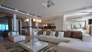 A modern living room with a large white sectional sofa, a glass coffee table, and a kitchen area in the background. - Beautiful 3 bedroom villa located within minutes of the beach and surrounded by green gardens (Gustavia)