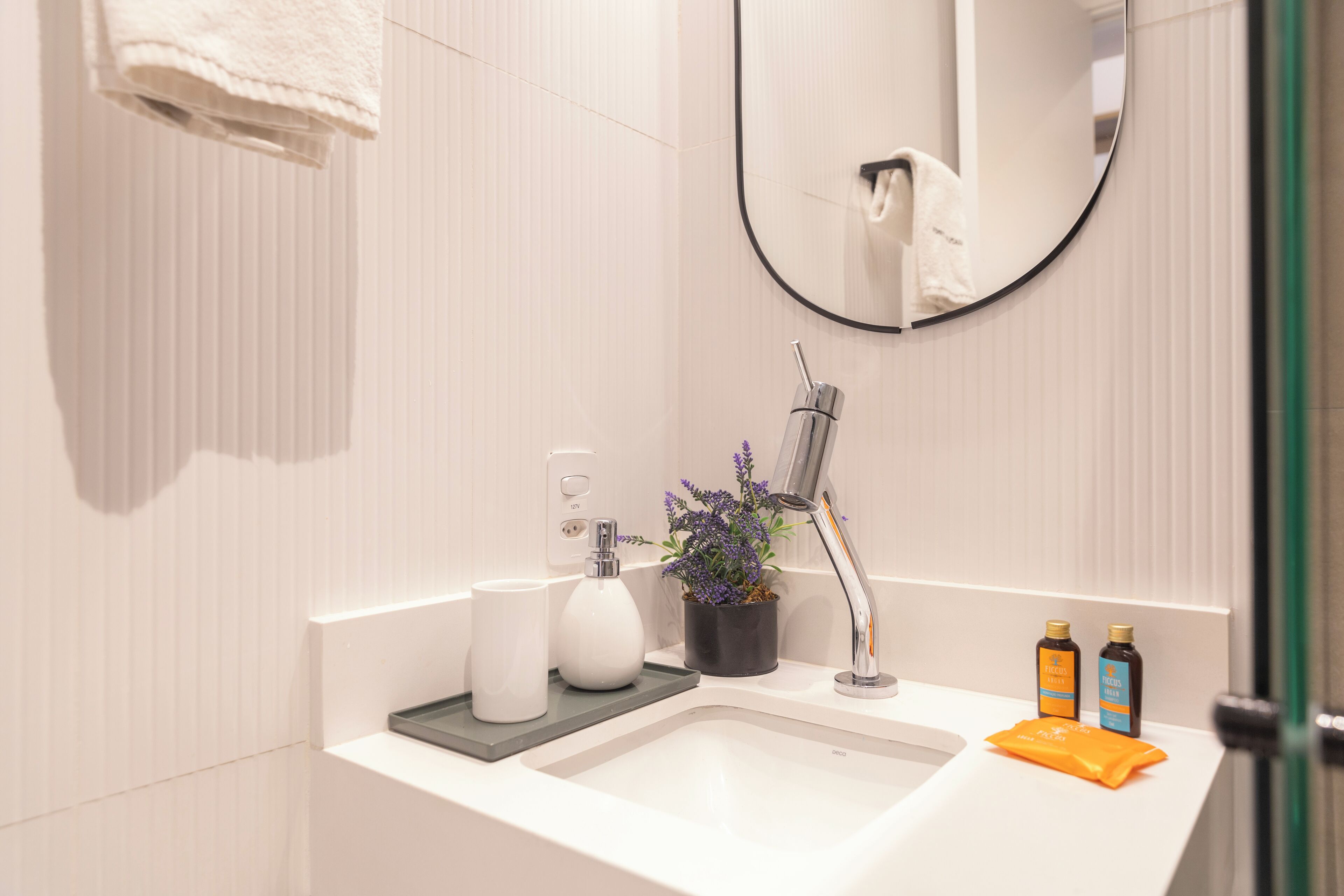 Un baño moderno con un lavabo blanco, un espejo, una toalla colgada en la pared, una planta en maceta y dos botellas de loción.
