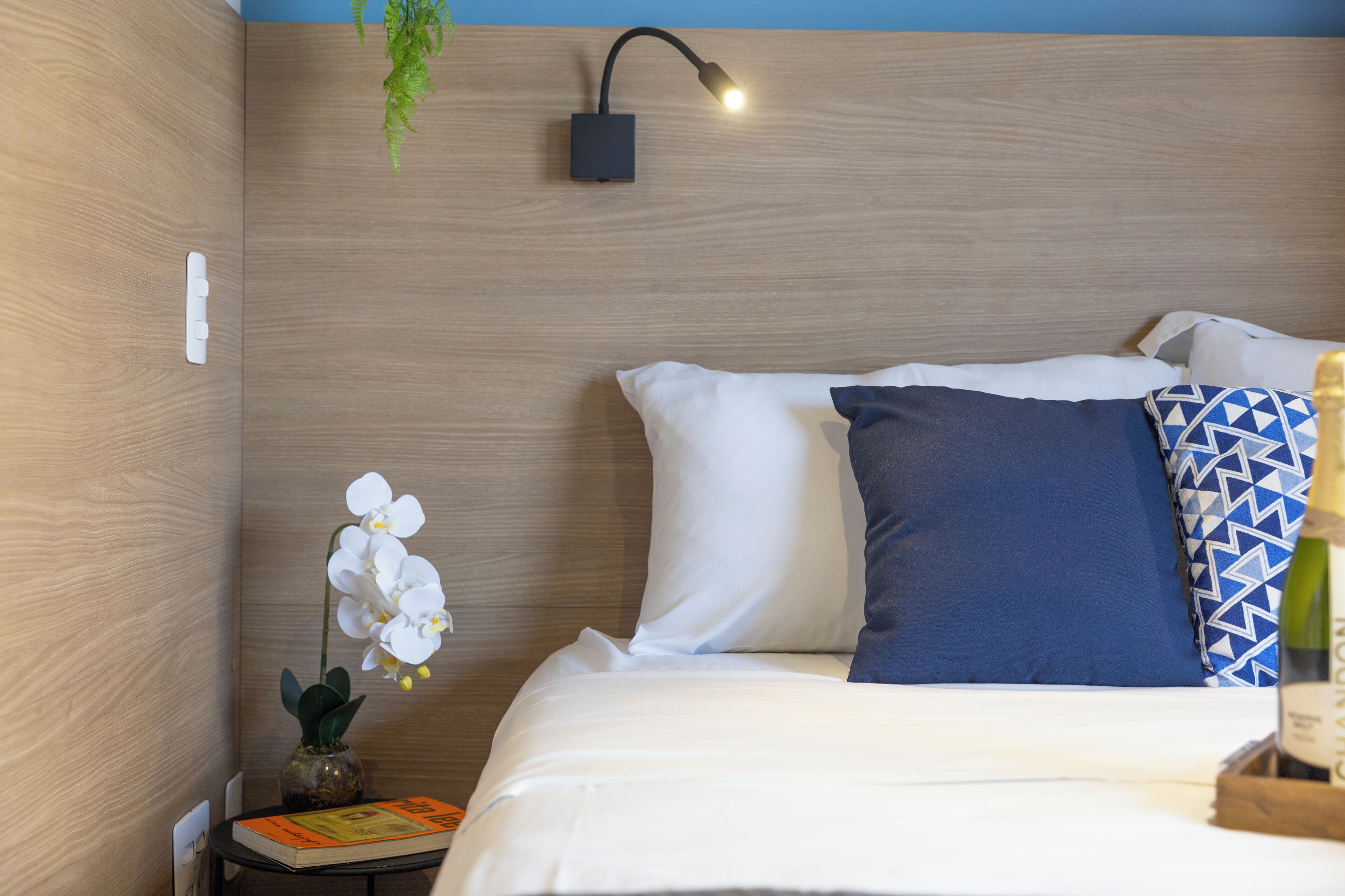 A neatly made bed with white and blue pillows, a small table with a book and a vase of flowers, and a wall-mounted light fixture.