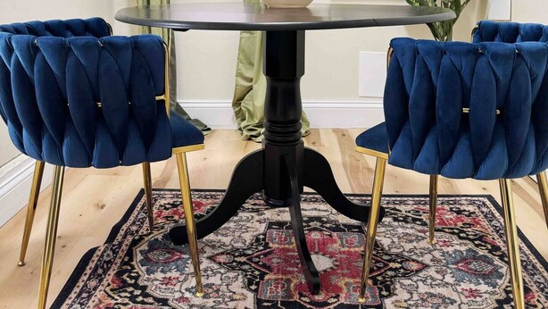 Comedor con mesa redonda, dos sillas tapizadas en azul, alfombra estampada, un jarrón con flores, un cuadro y una ventana con cortinas verdes.
