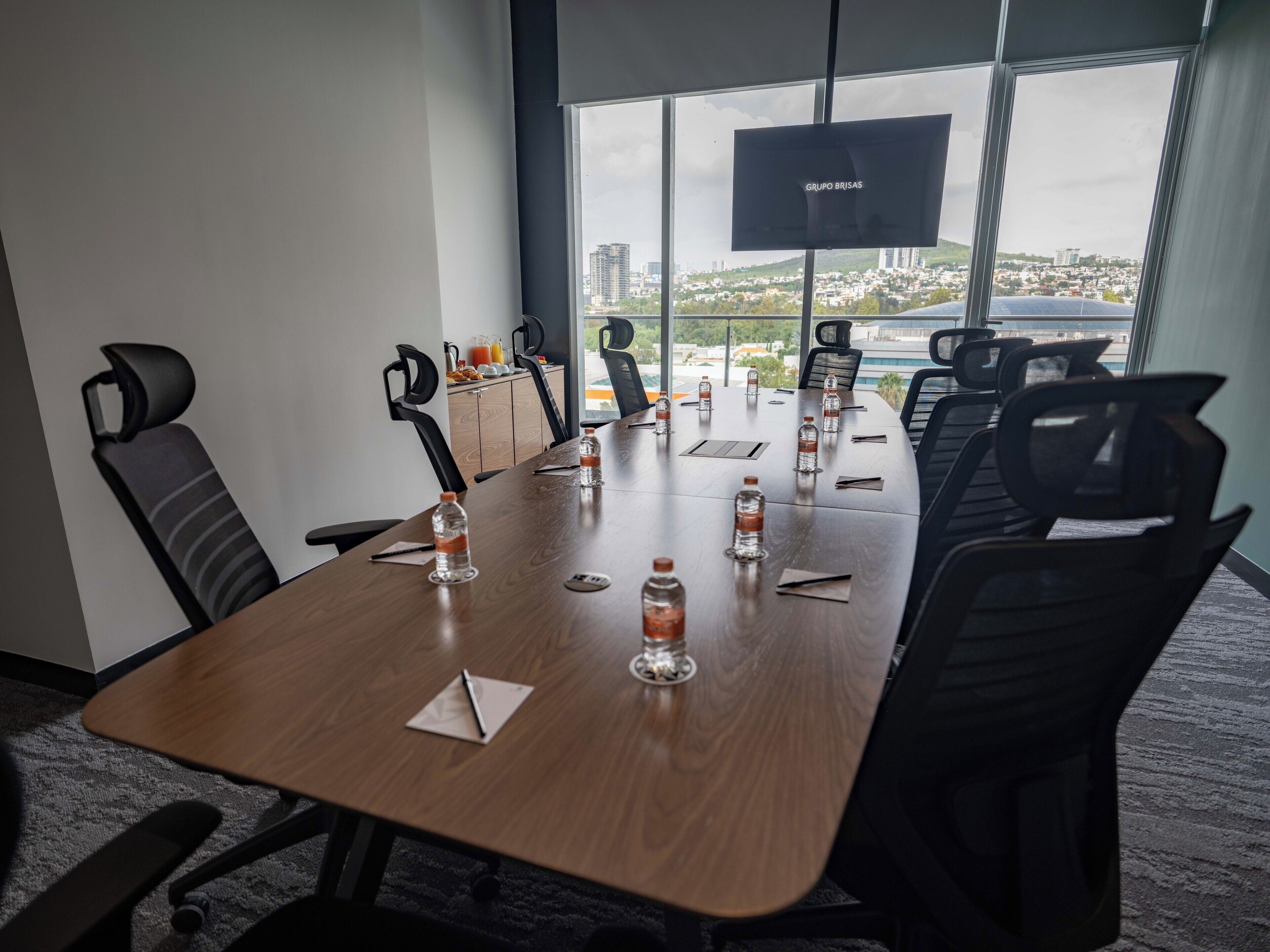 A conference room with a large table, chairs, a TV screen, and a view of a cityscape through large windows.
