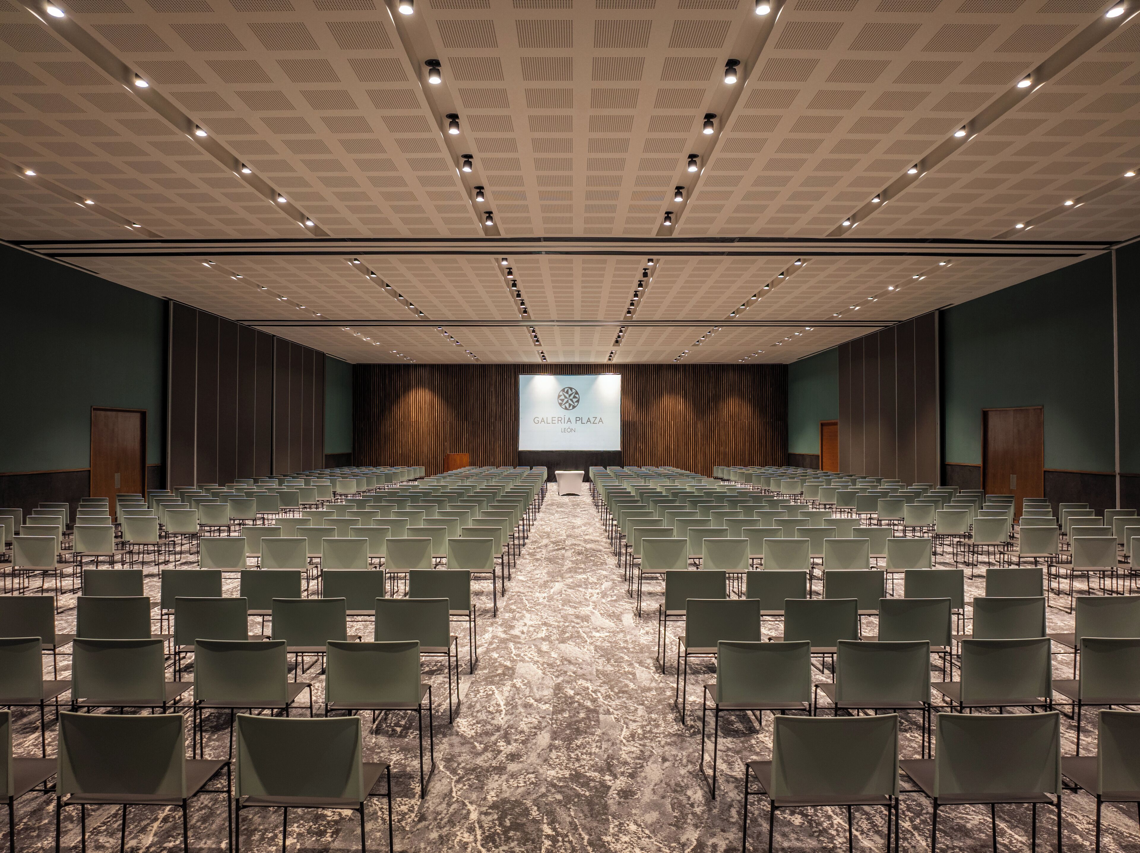 A conference room with rows of chairs, a large screen at the front, and a patterned ceiling.