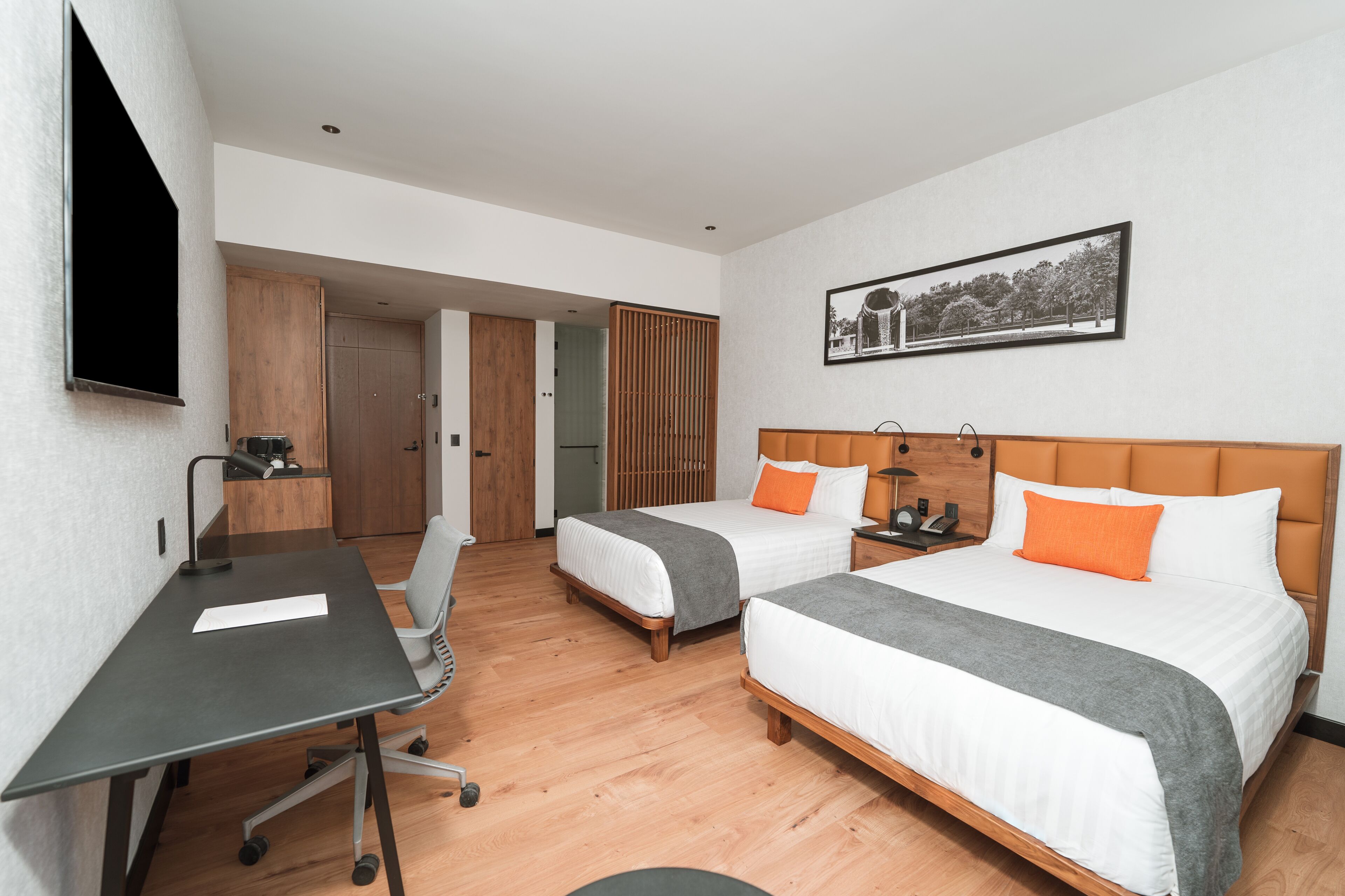 Quarto de hotel com duas camas, uma escrivaninha e uma cadeira. O chão é de madeira, há uma pintura na parede e uma porta que dá para outro cômodo.