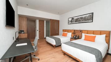 A hotel room with two beds, a desk, and a chair. There is a wooden floor, a painting on the wall, and a door leading to another room.