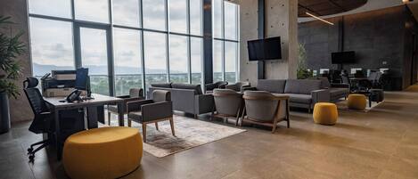 A modern office space with large windows, a sofa, chairs, a desk, and a television.
