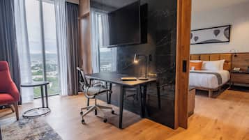 Et moderne hotelværelse med en stor seng, et skrivebord med stol, et TV ophængt på væggen og udsigt over byen gennem store vinduer.