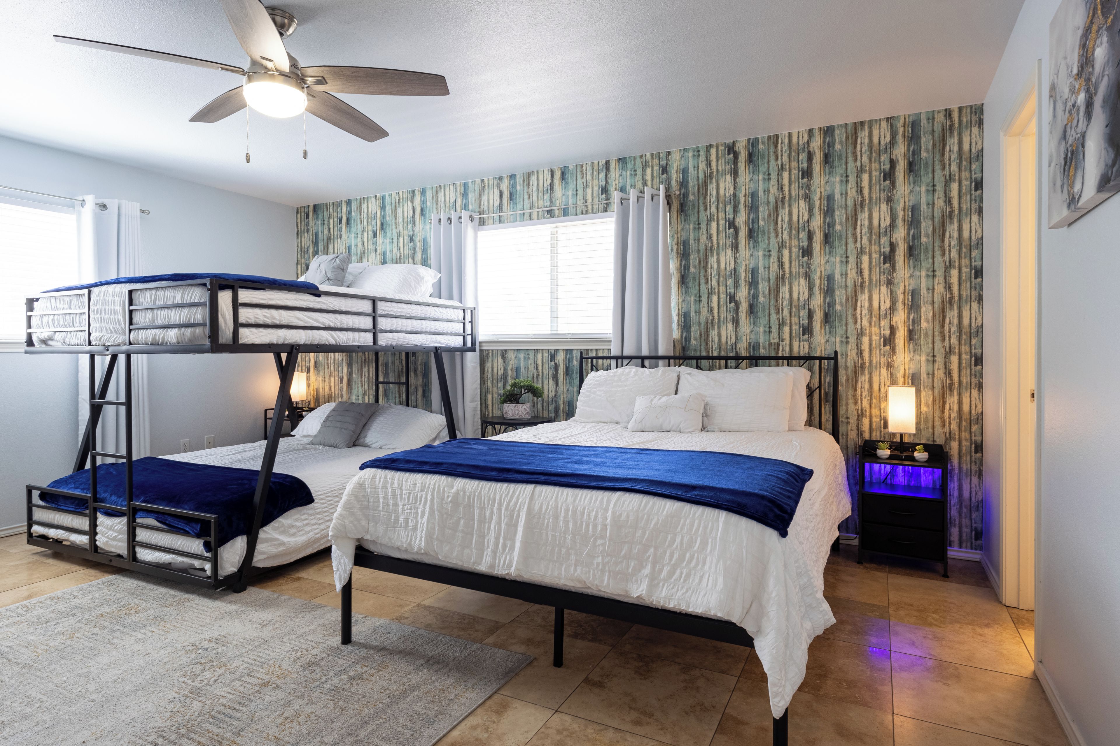 A room with bunk beds, a ceiling fan, a window with curtains, a nightstand with a lamp, and a wall with a textured pattern.