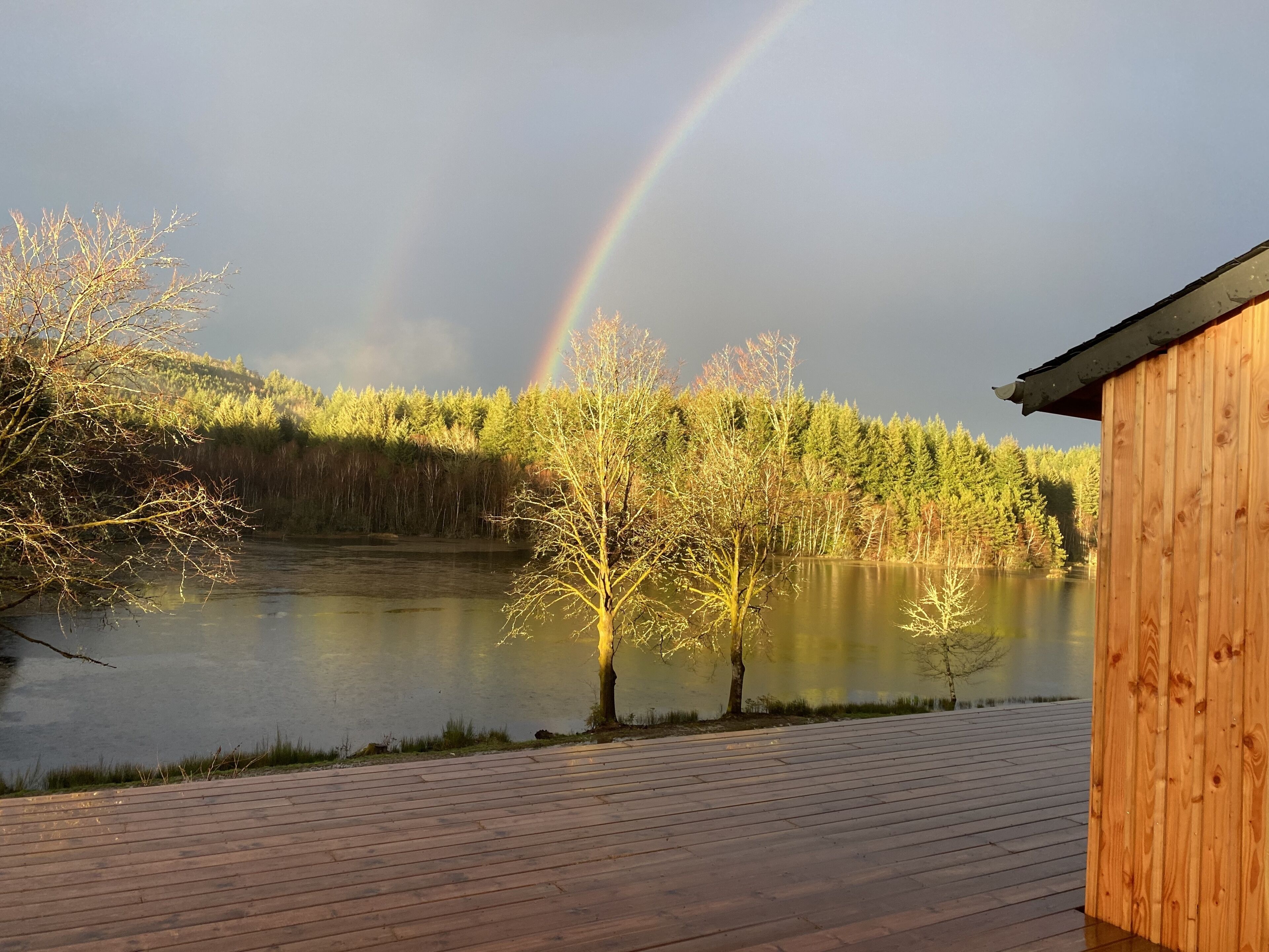 Eine Holzterrasse mit Blick auf einen stillen See, über dem ein Regenbogen erscheint, umgeben von Bäumen und einem bewaldeten Hang.