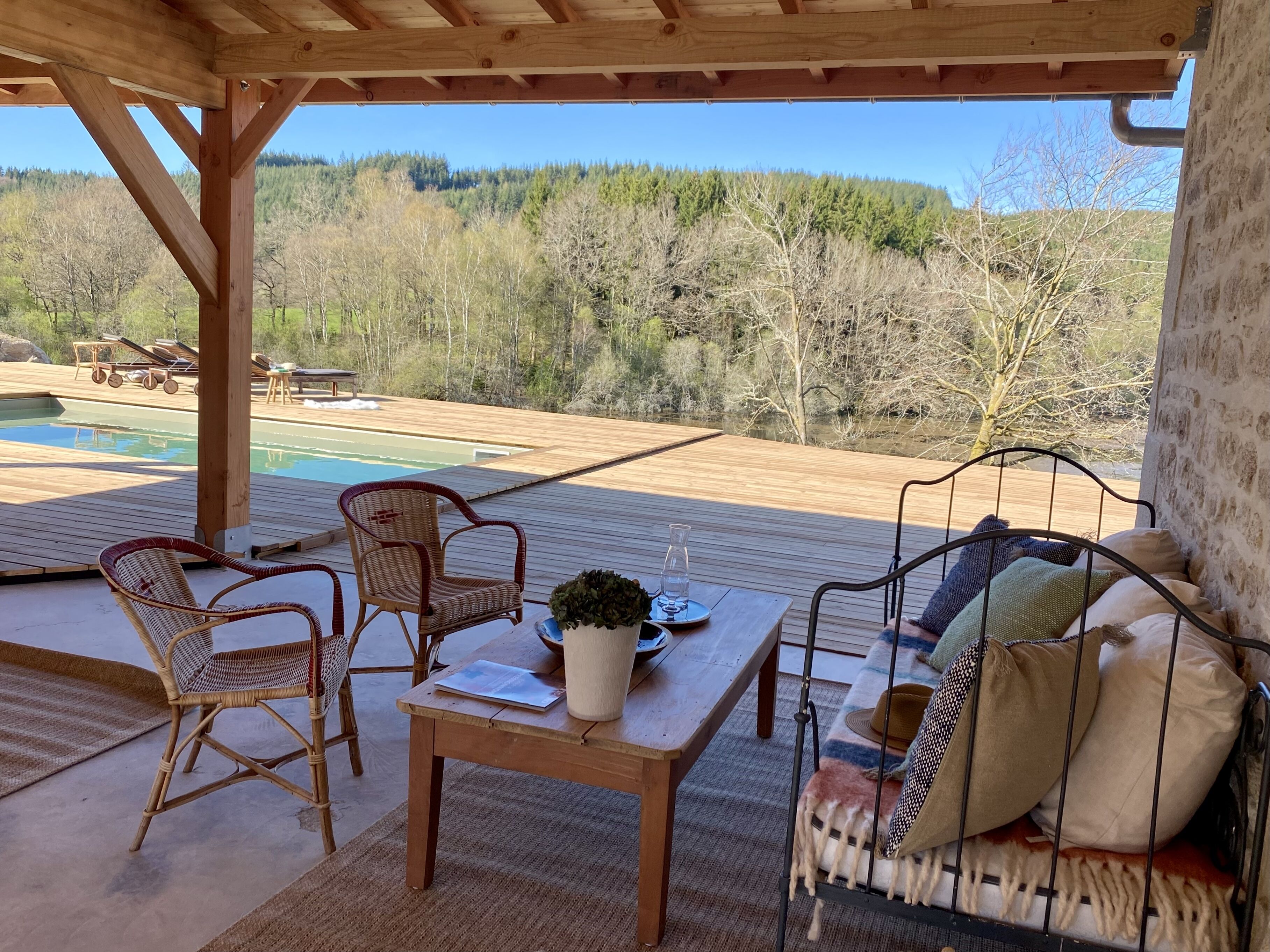 Une terrasse en bois avec des fauteuils en osier, une balançoire en métal et une vue sur une piscine et des collines boisées.
