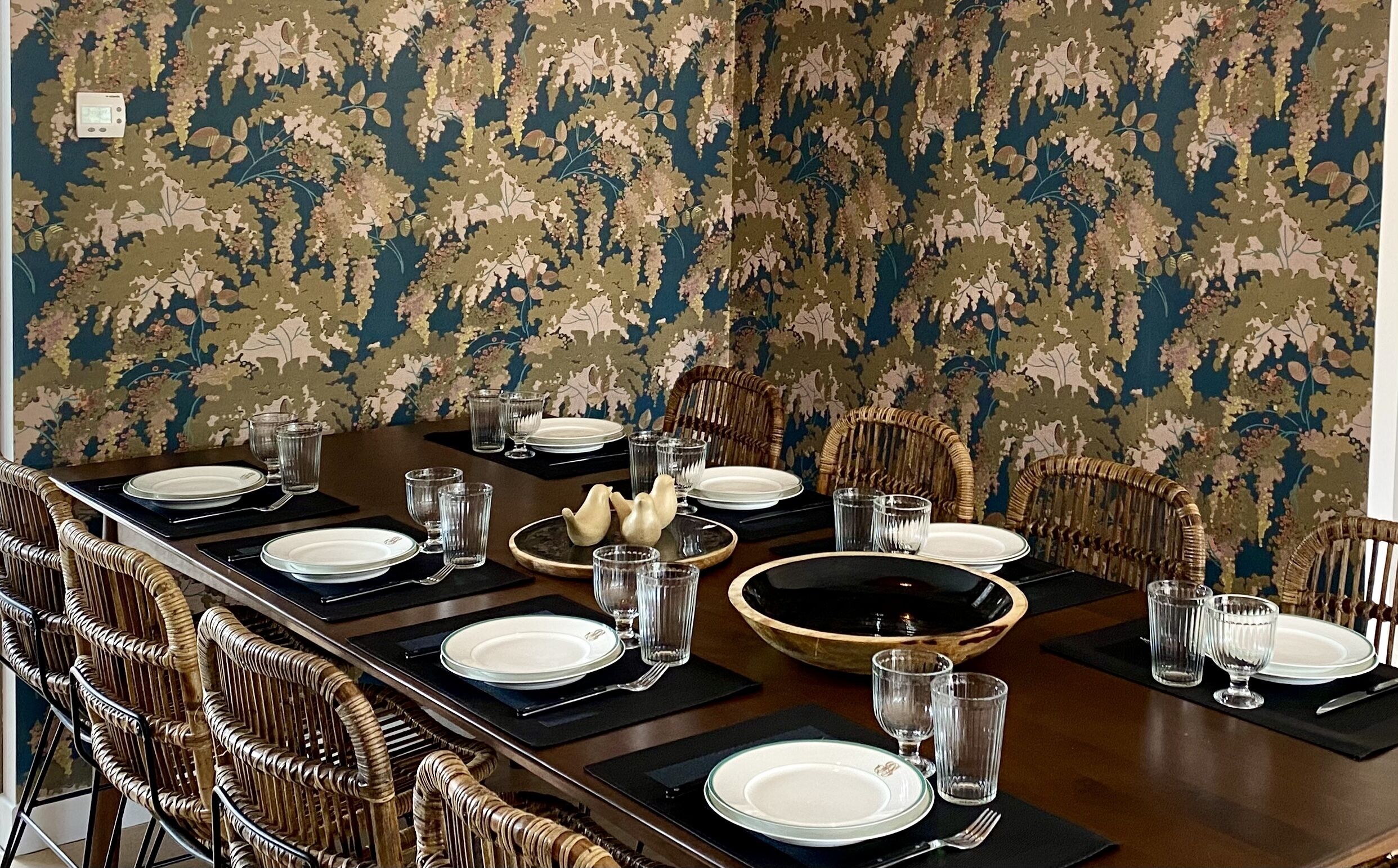 Une salle à manger avec une longue table dressée pour six personnes, entourée de chaises en osier.
