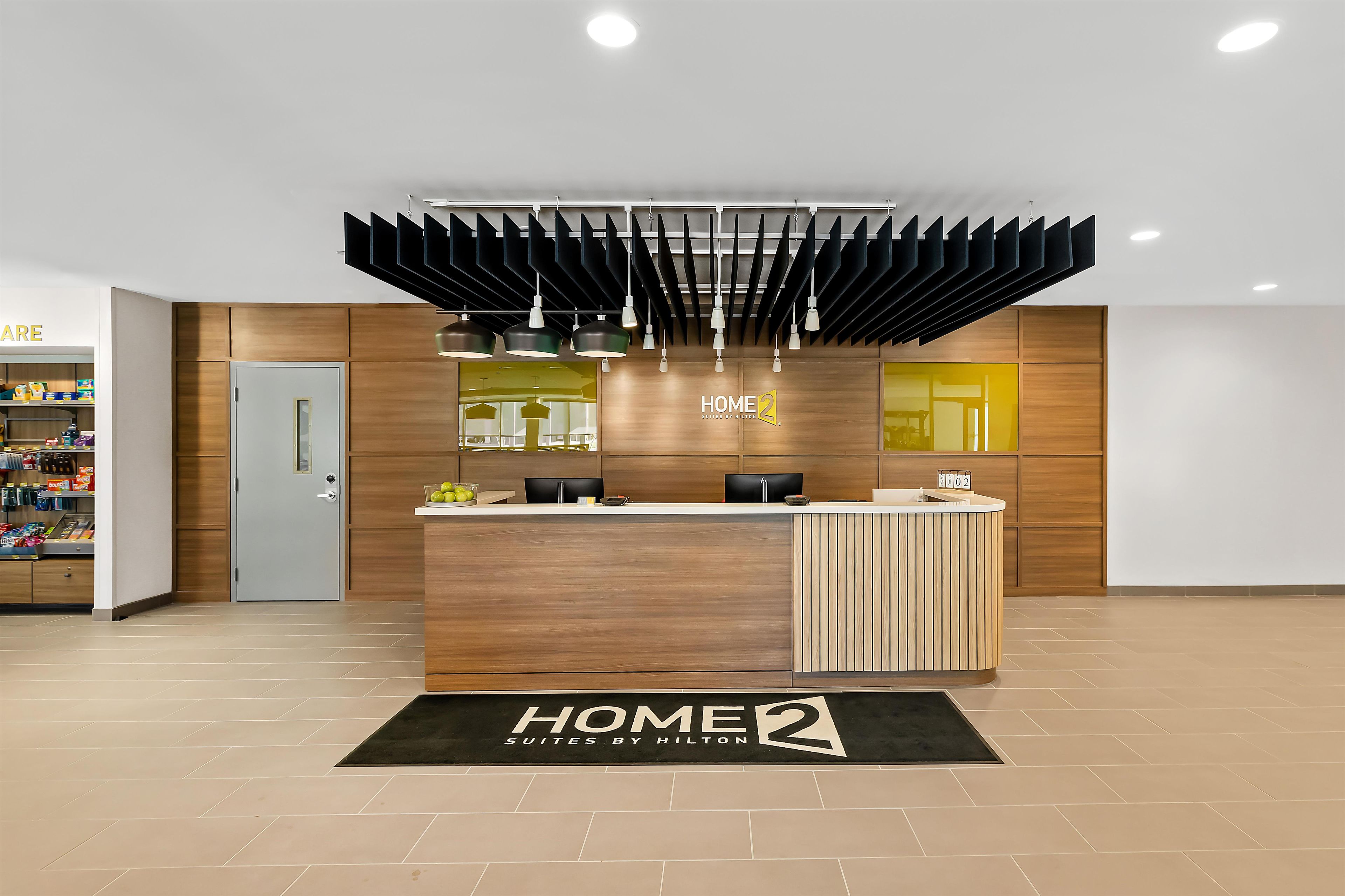 A hotel reception area with a modern design, featuring a reception desk, a mirror, and a sign that reads 'HOME 2 SUITES BY HILTON'.
