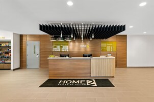 Un área de recepción de hotel con diseño moderno, que incluye un mostrador de recepción, un espejo y un cartel que dice 'HOME 2 SUITES BY HILTON'.