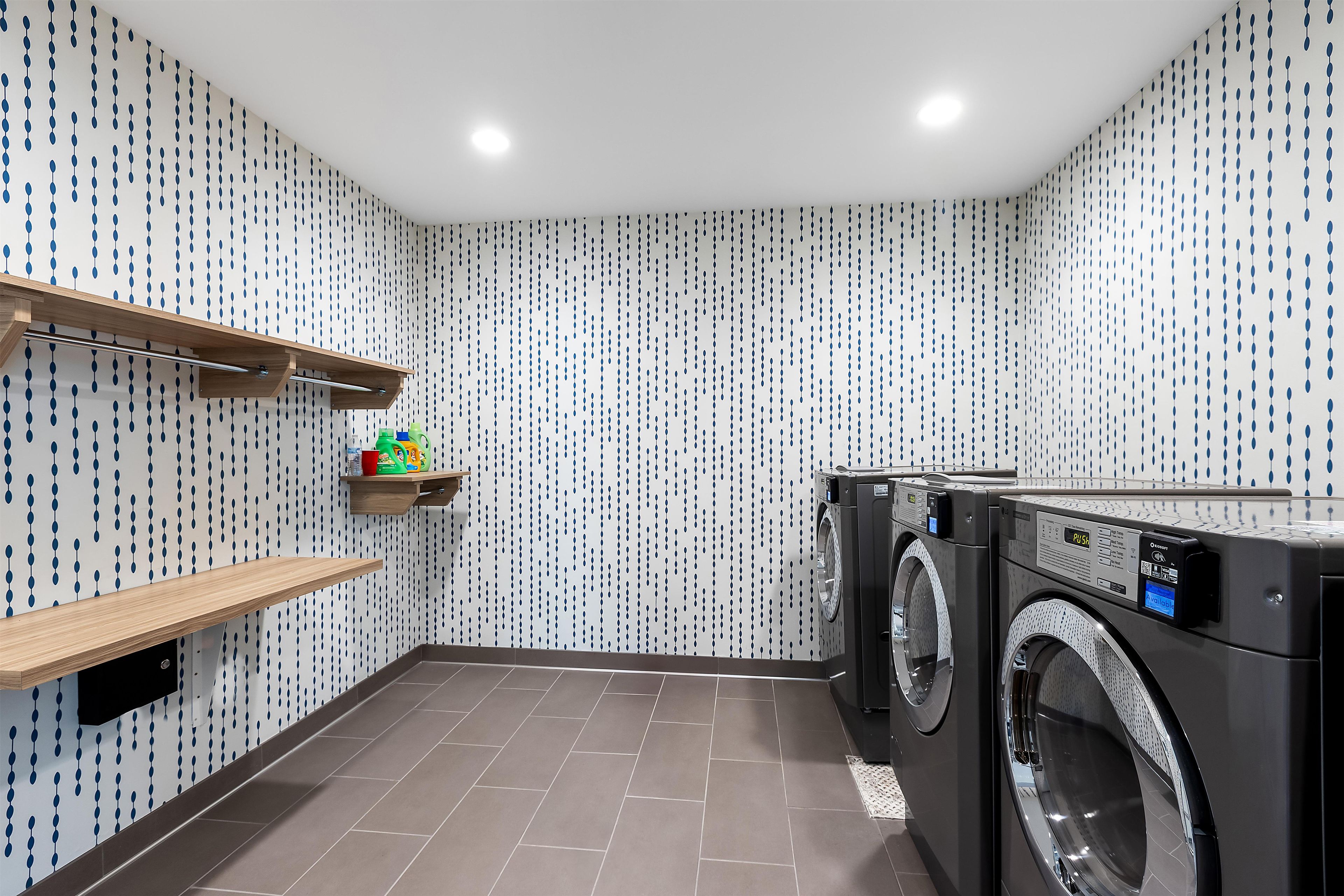 Une buanderie avec un papier peint à motifs, une étagère en bois contenant des produits de lessive et deux machines à laver à chargement frontal.