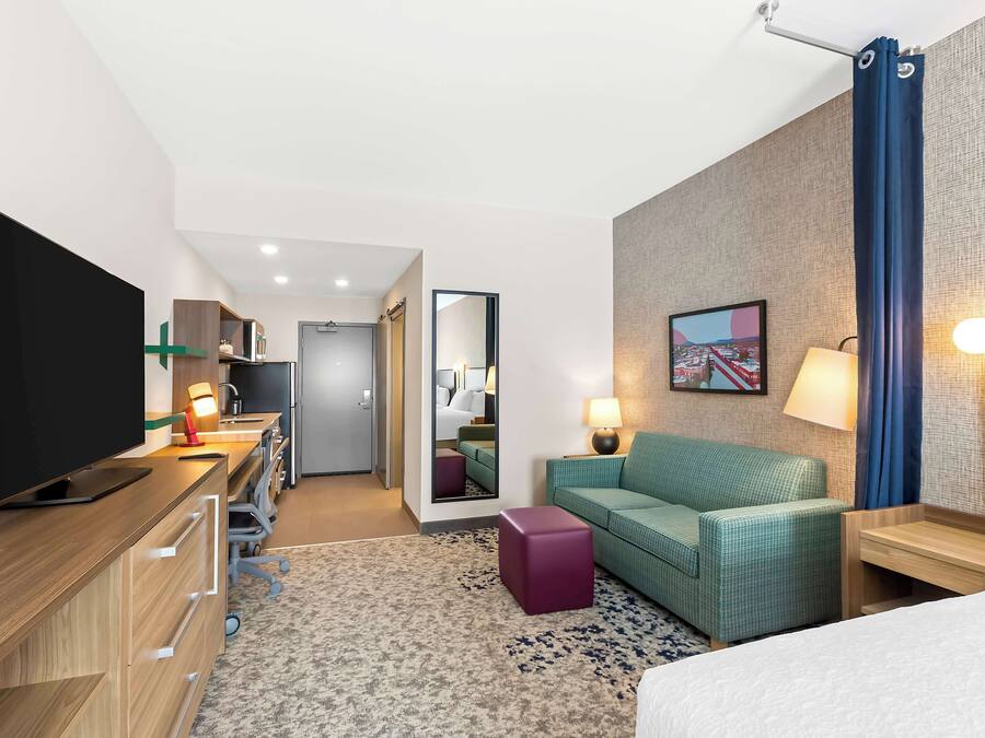 A hotel room with a bed, a desk with a flat-screen TV, a green sofa, a small table, a mirror, and a lamp.