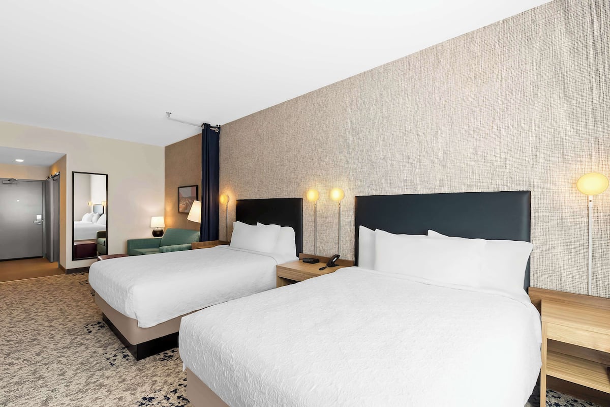 Habitación de hotel con dos camas, una mesita de noche de madera, un espejo y una pared con papel tapiz texturizado.