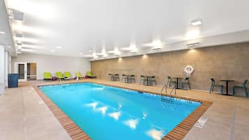 An indoor swimming pool with clear lane markings, surrounded by green chairs and tables, and a tiled floor.
