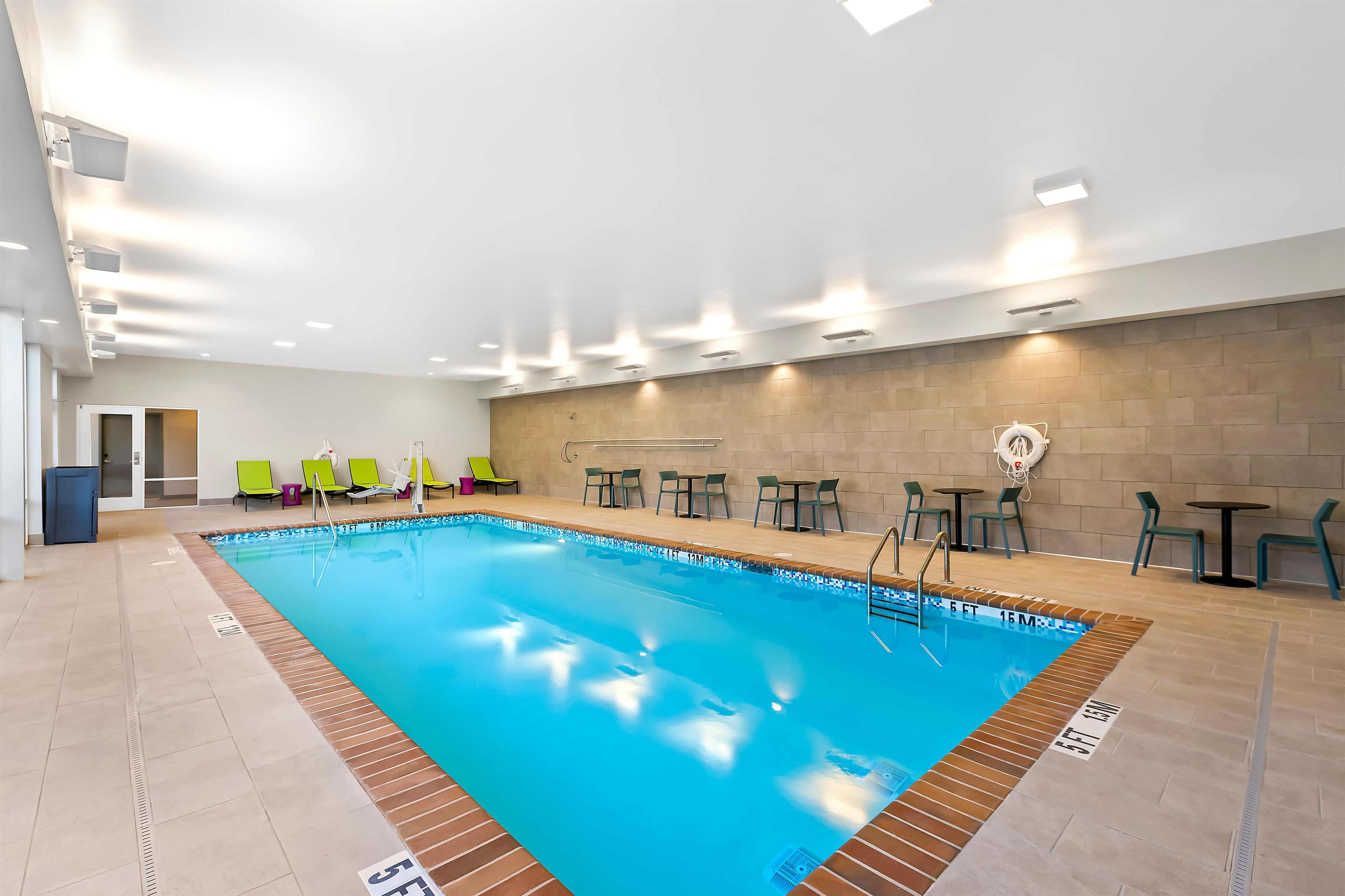 An indoor swimming pool with clear lane markings, adjacent lounge chairs, and tables.