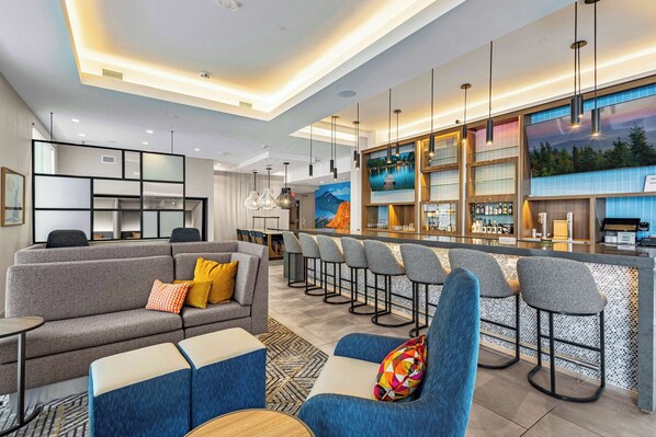 A modern hotel lobby with a bar area, seating arrangements, and a kitchen visible in the background.