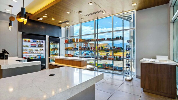 Un espacio comercial moderno con mostrador de mármol, vitrina refrigerada y vista al estacionamiento a través de amplios ventanales.
