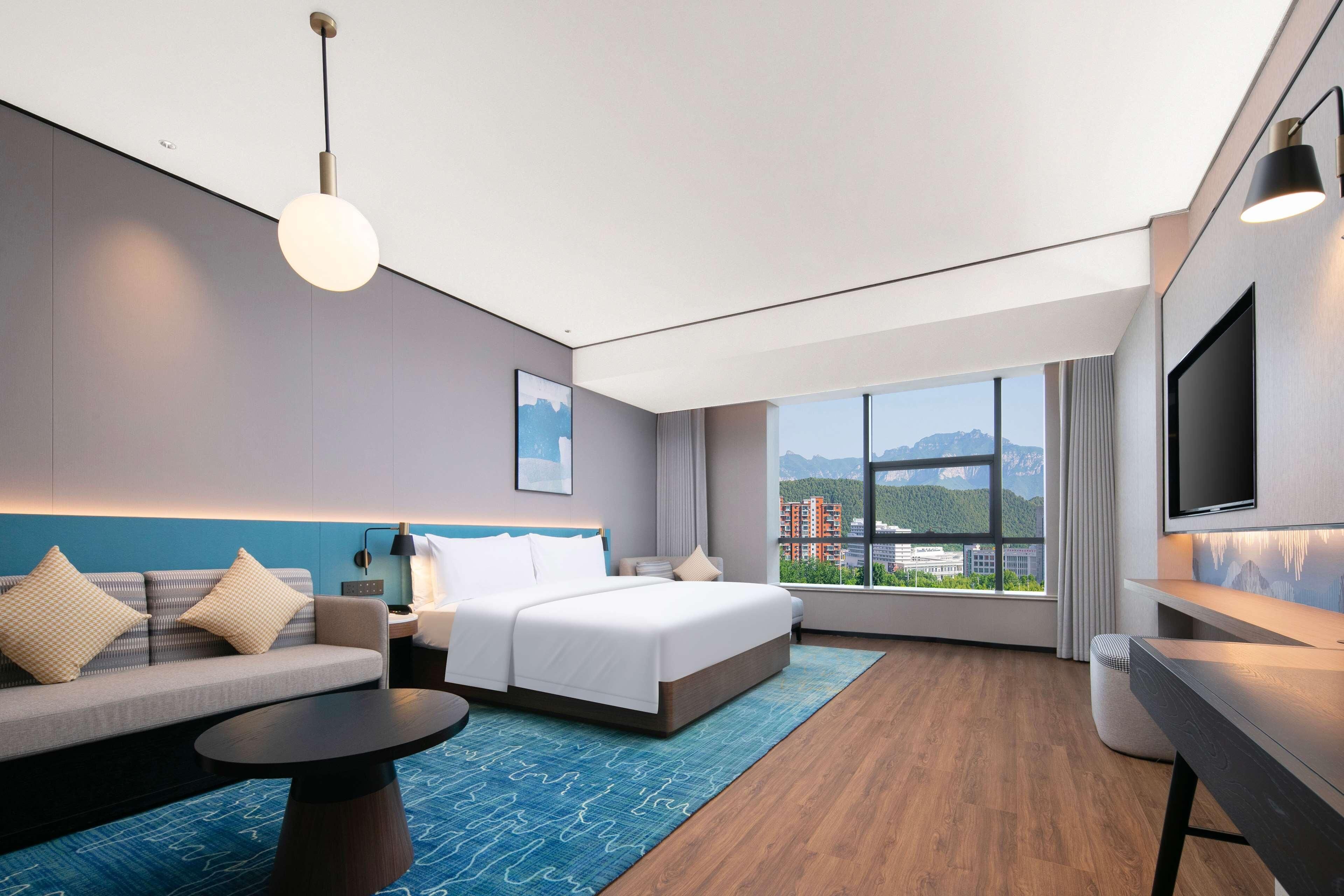 A modern hotel room with a bed, sofa, desk, and a large window offering a city view.