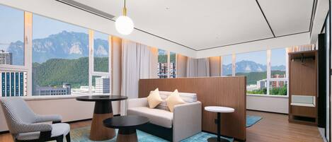 A modern hotel room with a large window offering a mountain view, a sofa, a chair, and a small table.