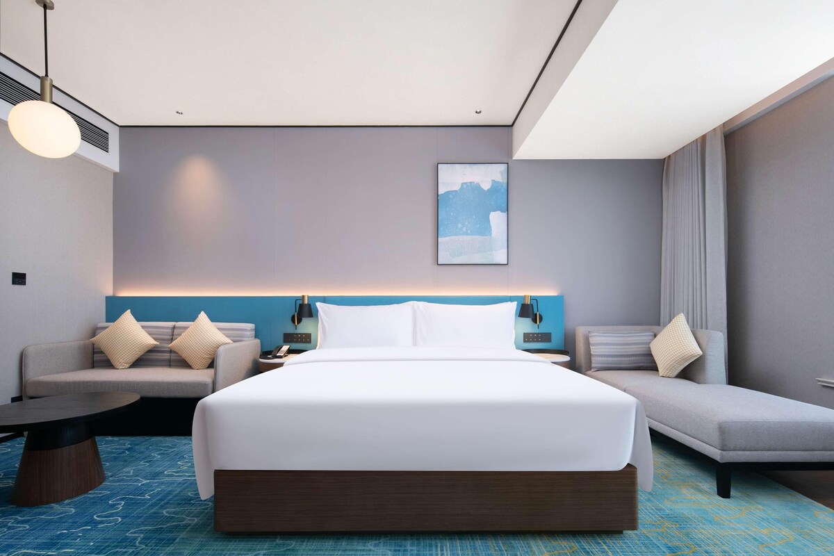 A modern hotel room with a large bed, a sofa, a desk, and a wall-mounted artwork.