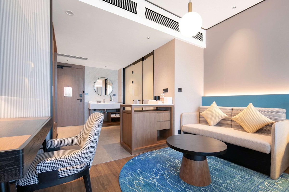 A modern hotel room with a sofa, a round coffee table, a houndstooth patterned chair, a small kitchen area, and a large mirror.