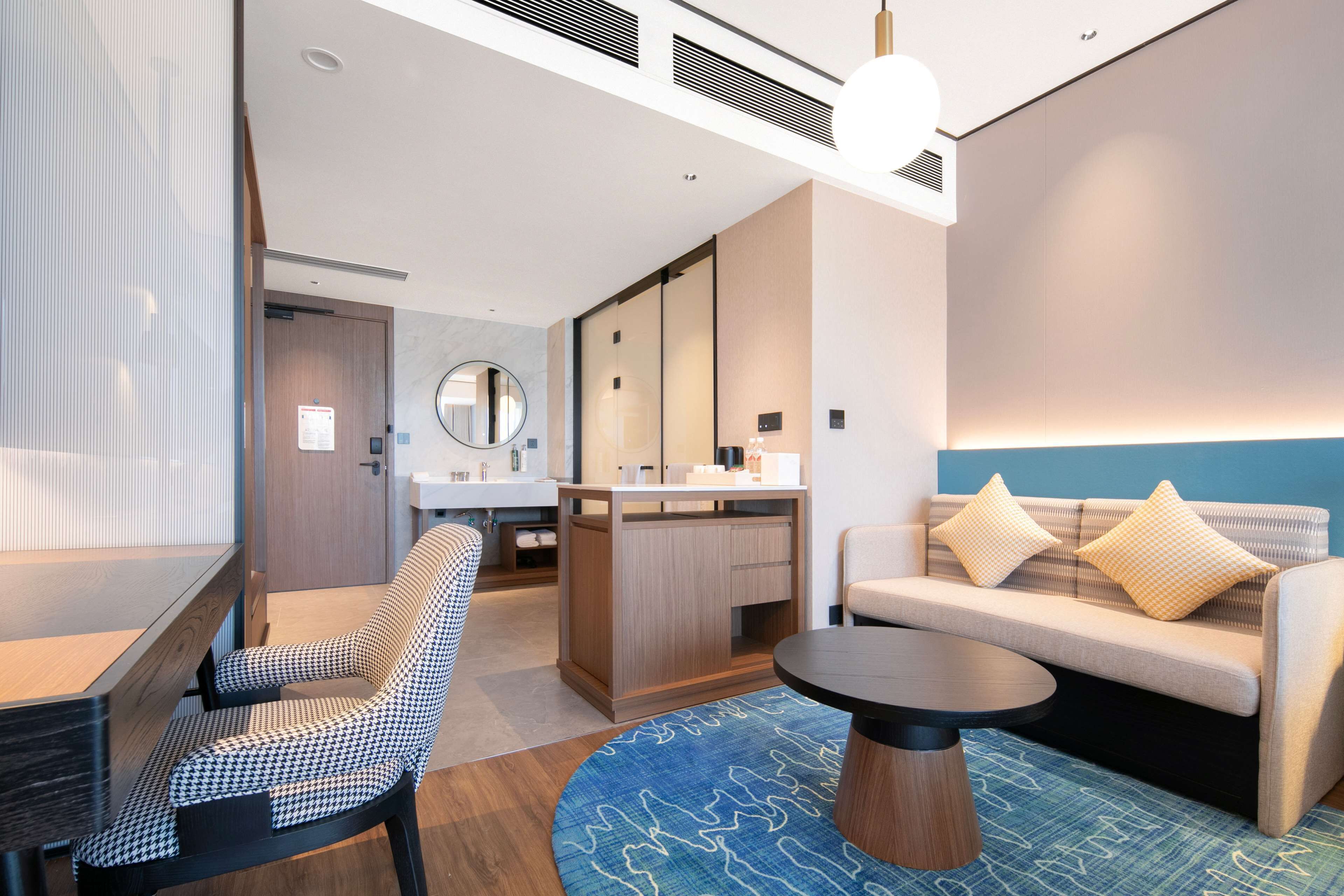 Una habitación de hotel moderna con sofá, una mesa de centro redonda, una butaca estampada de pata de gallo, un mueble de madera, un espejo y una alfombra azul estampada.