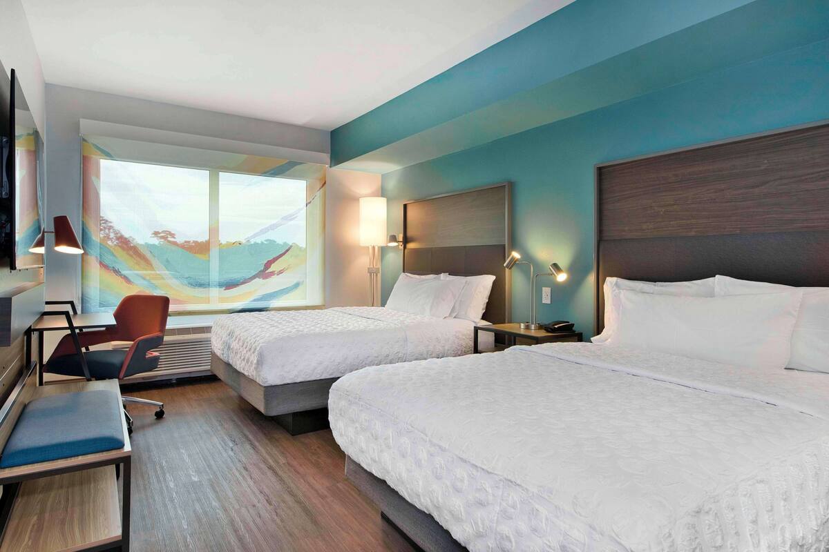 Une chambre d’hôtel avec deux lits, un bureau, une chaise et une grande fenêtre donnant sur un paysage coloré.