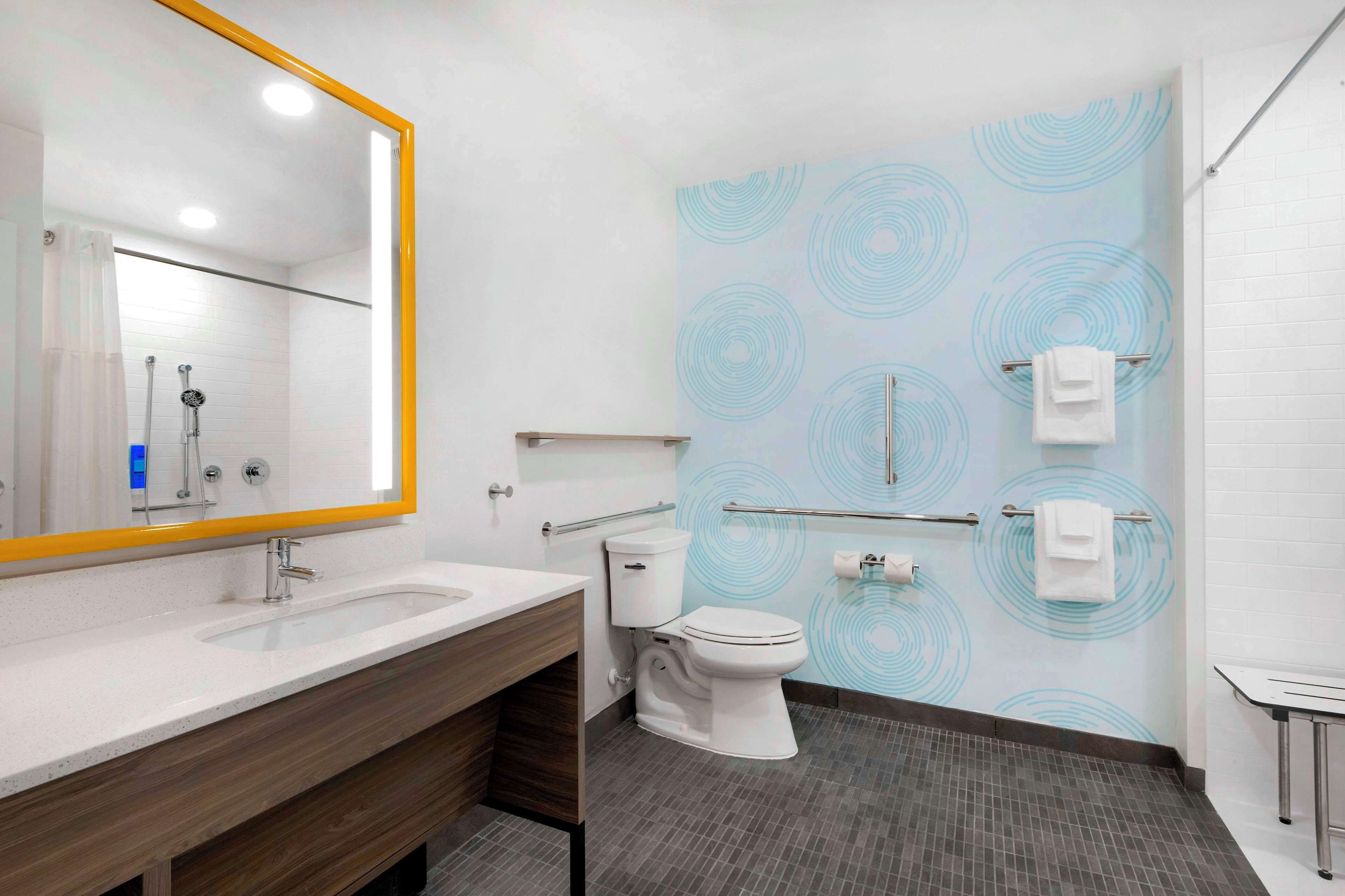 A modern bathroom with a large mirror, white sink, and toilet. There are towels hanging on a rack and a shower area visible in the background.