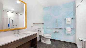 A modern bathroom with a large mirror, white sink, and toilet. There are towels hanging on a rack and a shower area visible in the background.
