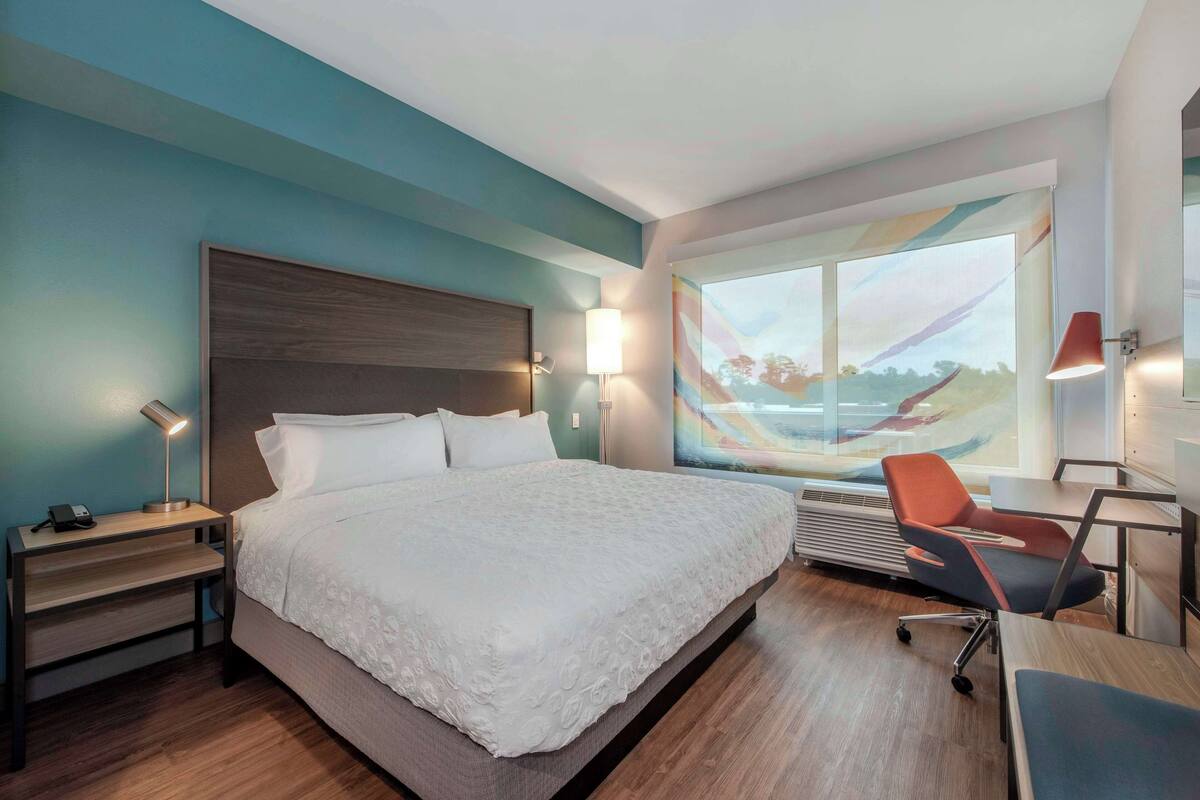 Une chambre d’hôtel moderne, dotée d’un grand lit, d’un bureau et d’une chaise. Une fenêtre offre une vue sur le paysage.
