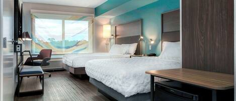 A hotel room with two beds, a desk, a chair, a TV, and a view of the beach.
