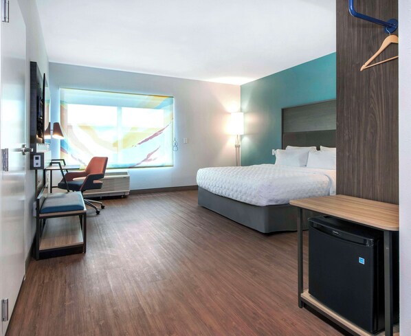 A modern hotel room with a bed, desk, chair, and a small refrigerator.