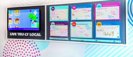 Un mur sur lequel est accrochée une télévision diffusant un match, ainsi que divers panneaux avec des notes manuscrites sur les attractions locales, les restaurants et les magasins.