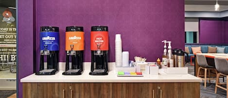 A coffee station with three dispensers labeled 'Lavazza' and 'Hot Water' in a modern room with purple walls and wooden cabinets.