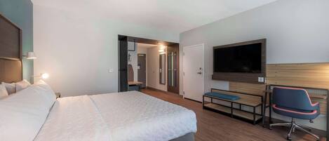 A modern hotel room with a large bed, a desk with a chair, a flat-screen TV, and a walk-in closet.