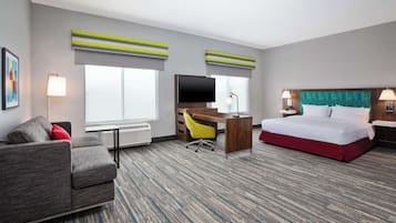 A hotel room with a grey sofa, a bed with a red accent, a desk with a TV, and two windows with blinds.