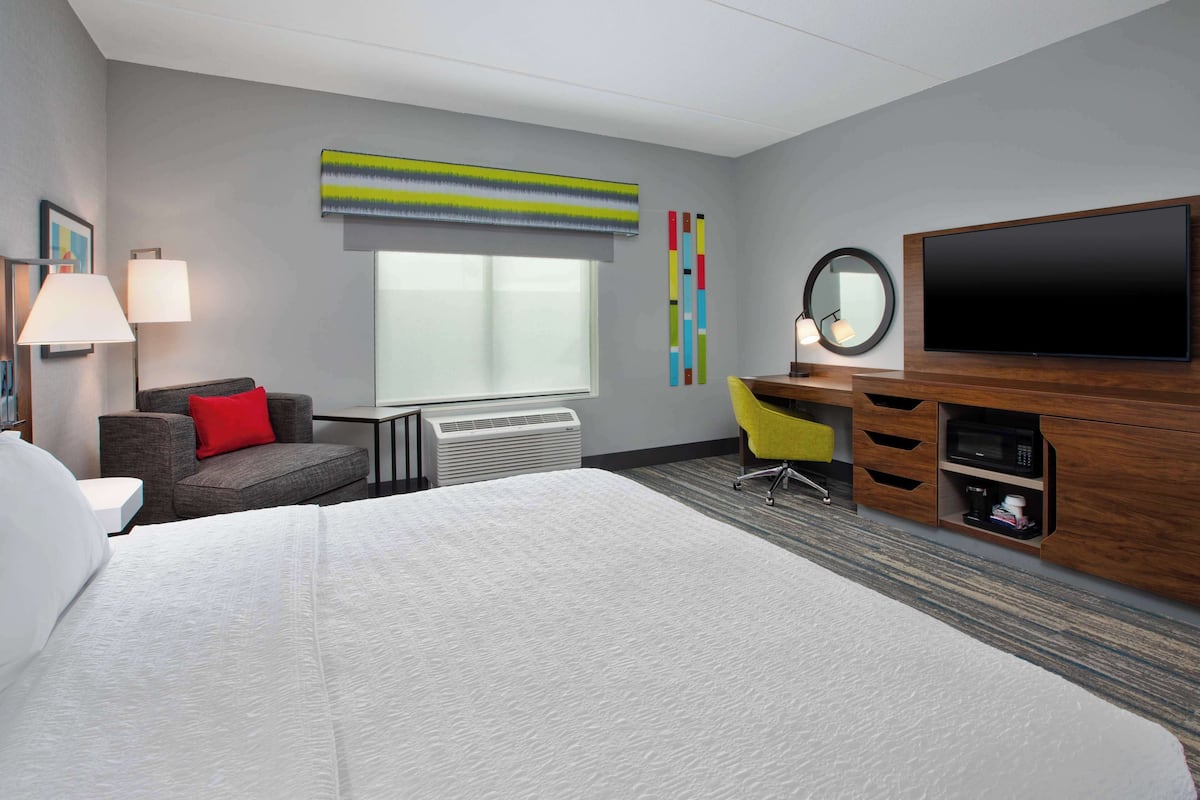 Une chambre d’hôtel avec un grand lit, un bureau doté d’un téléviseur à écran plat, une chaise, une lampe et une fenêtre avec des stores.