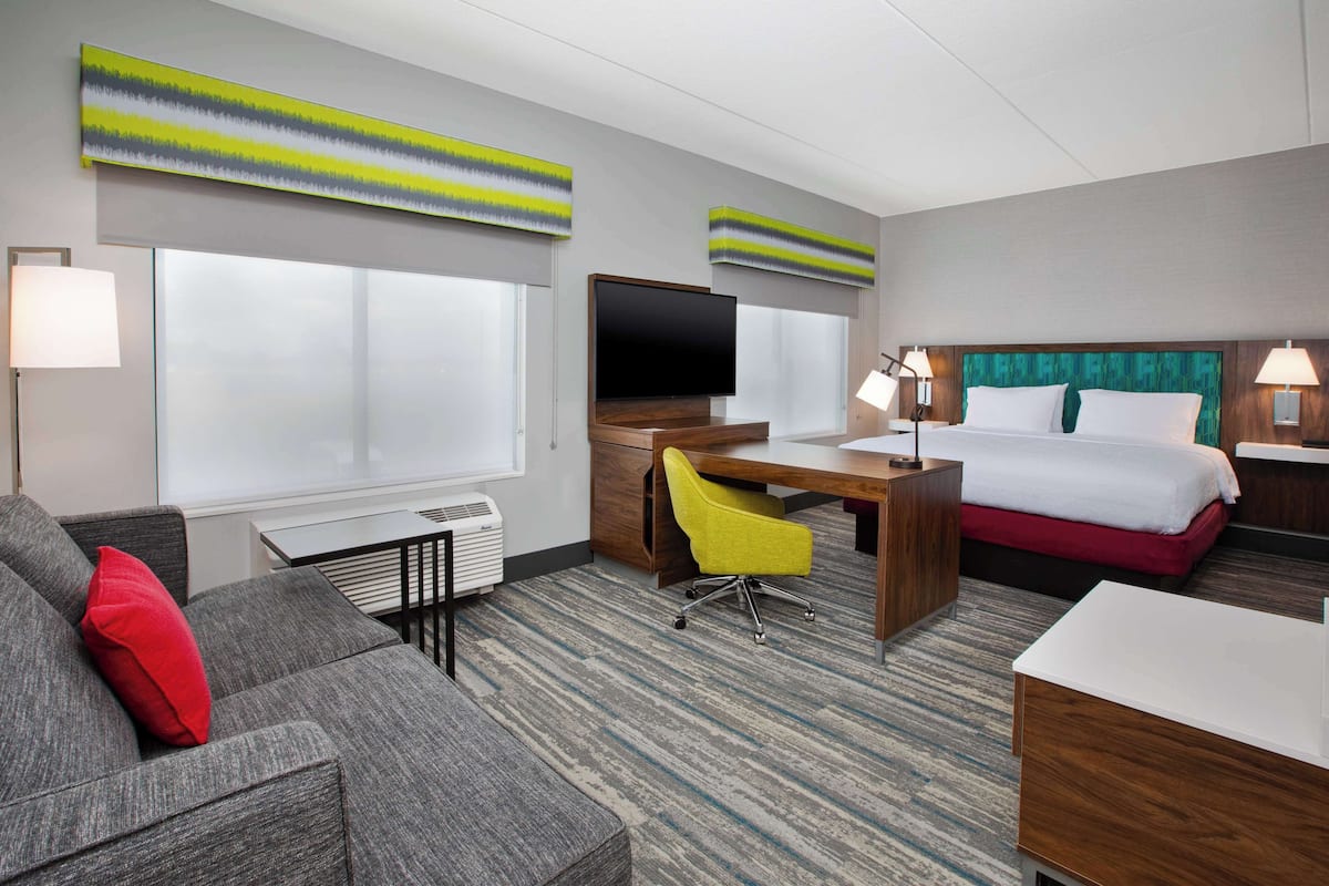 Une chambre d’hôtel avec un canapé gris, un fauteuil jaune, un bureau avec un téléviseur à écran plat, un lit avec une tête de lit verte et des œuvres d’art accrochées aux murs.