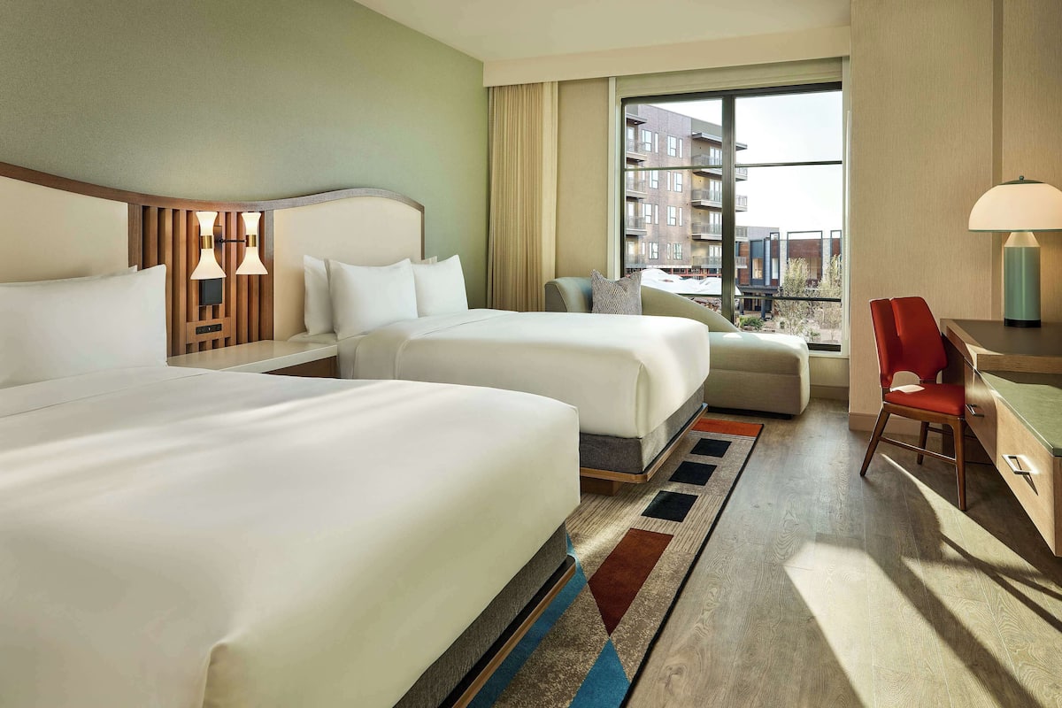 A hotel room with two beds, a desk, a chair, and a large window with a view of buildings.